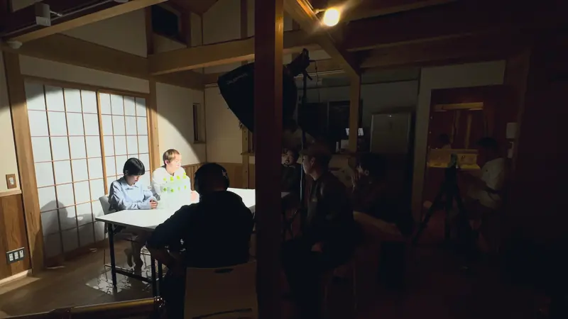 和室のセットで、照明の当たる机に出演者が座り、手前の暗がりでスタッフが撮影の準備をしている様子